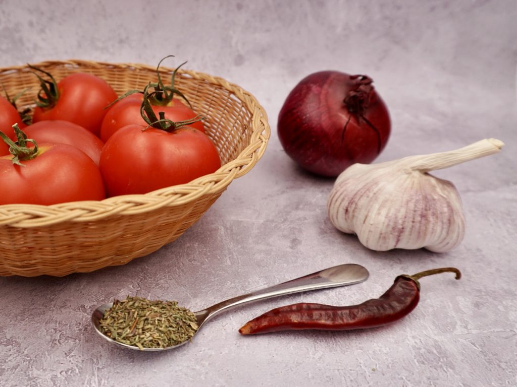 A basket of tomatoes, an onion, bulb of garlic, a chilli and a spoonful of dried herbs.
