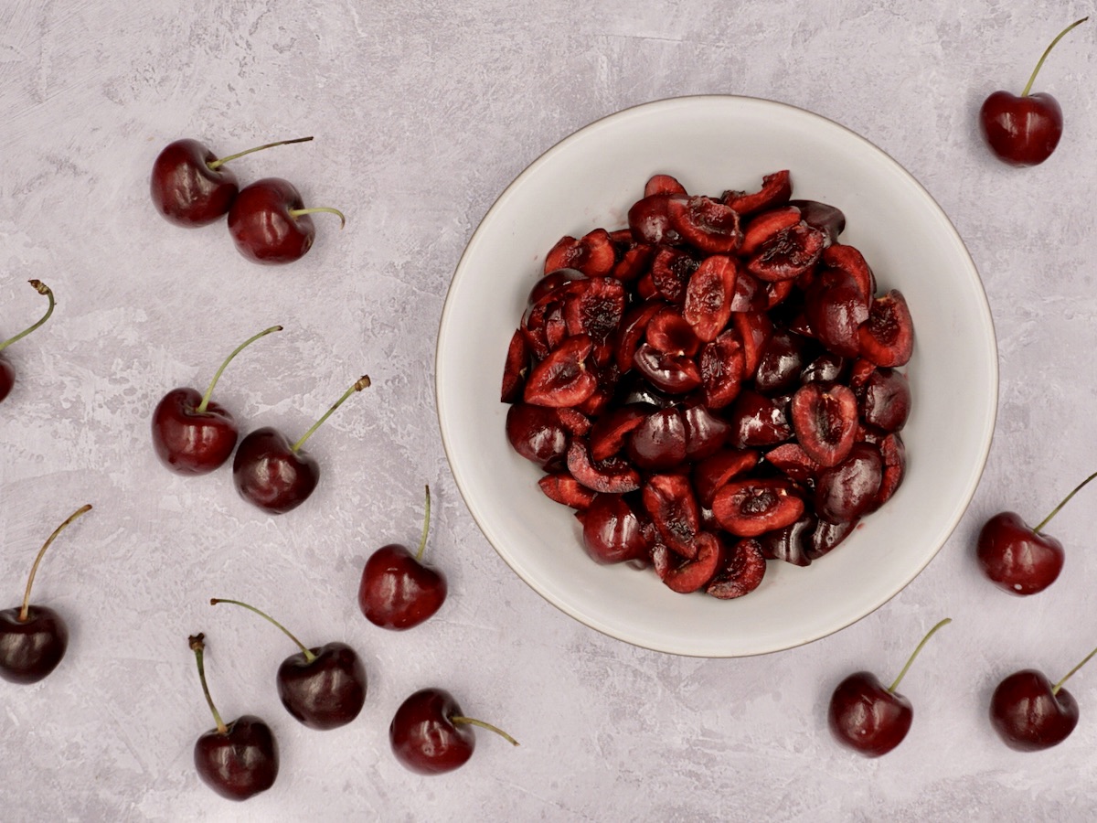 Stoned and halved cherries in a bowl next to whole fresh cherries