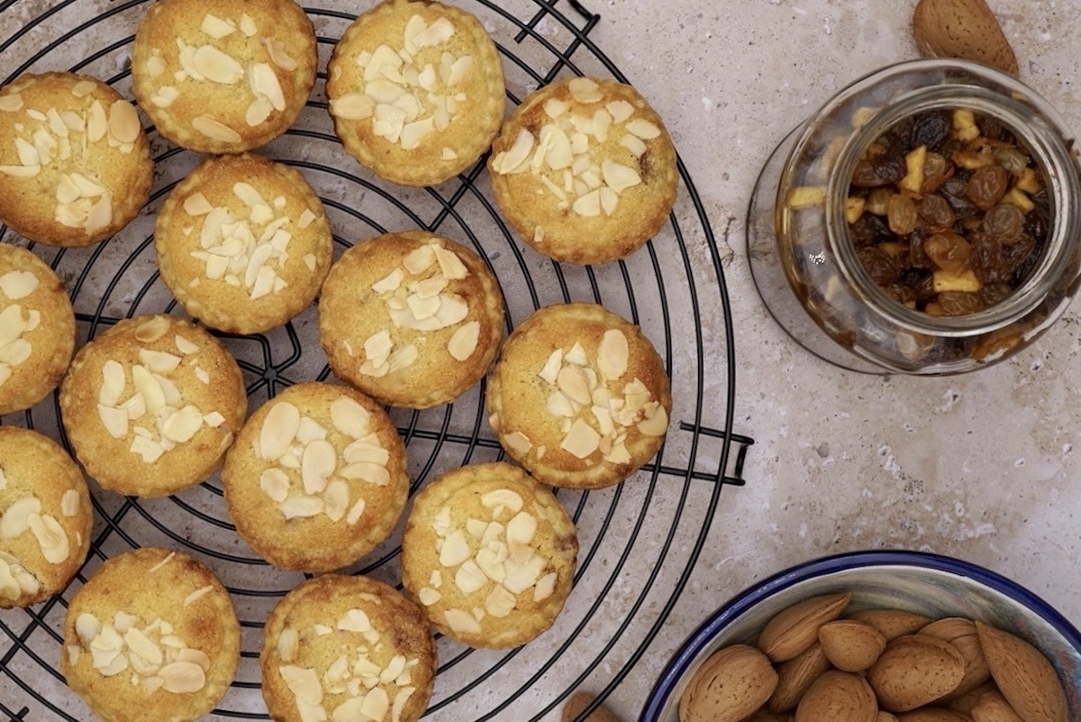 Overhead view of frangipane mince pies on a wire rack next to a jar of mincemeat and some whole almonds.