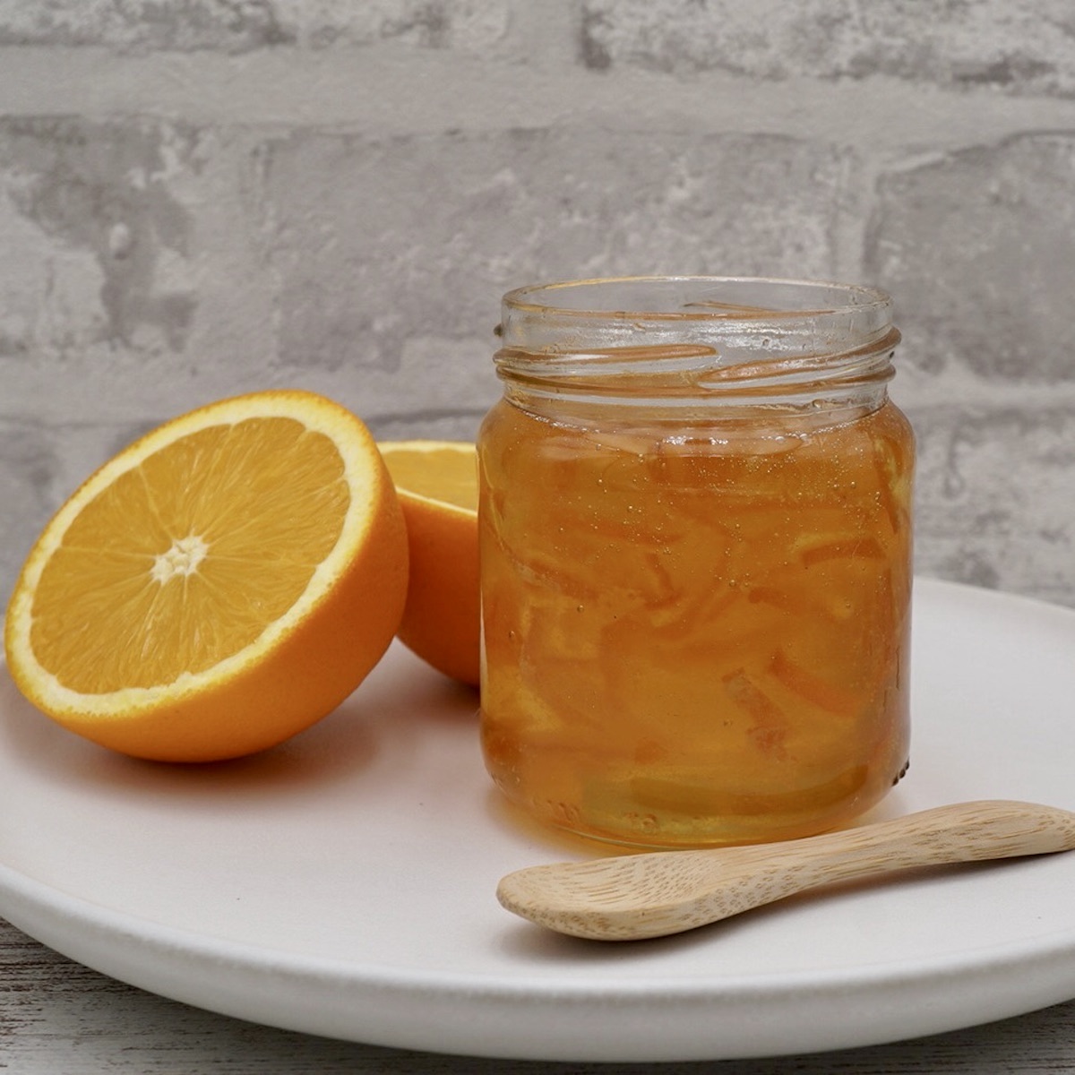 An open jar of Seville orange marmalade on a plate next to a spoon and an orange cut in half.