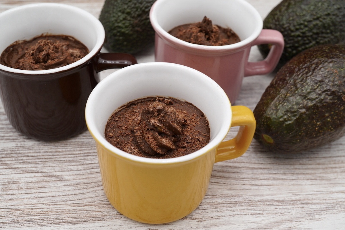 Three small cups containing chocolate mousse next to some avocados. 