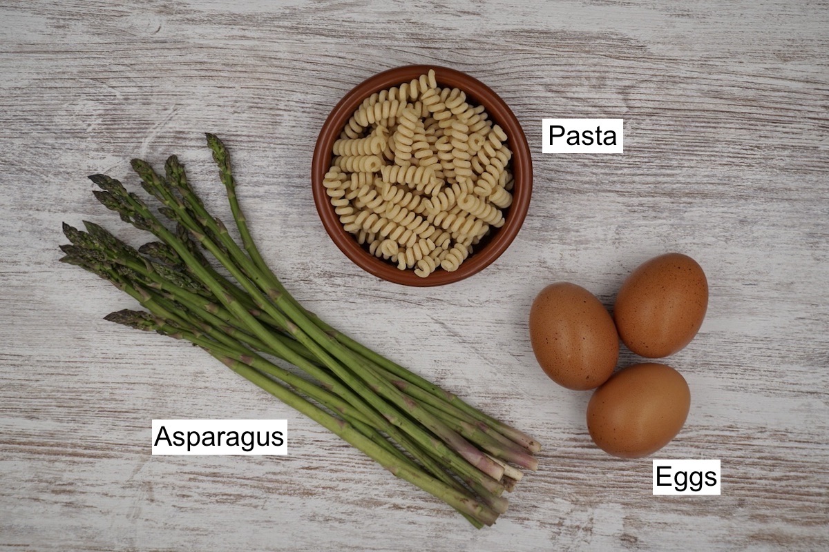Labelled cooking ingredients including a bowl of dried pasta, a bunch of fresh asparagus and three eggs.