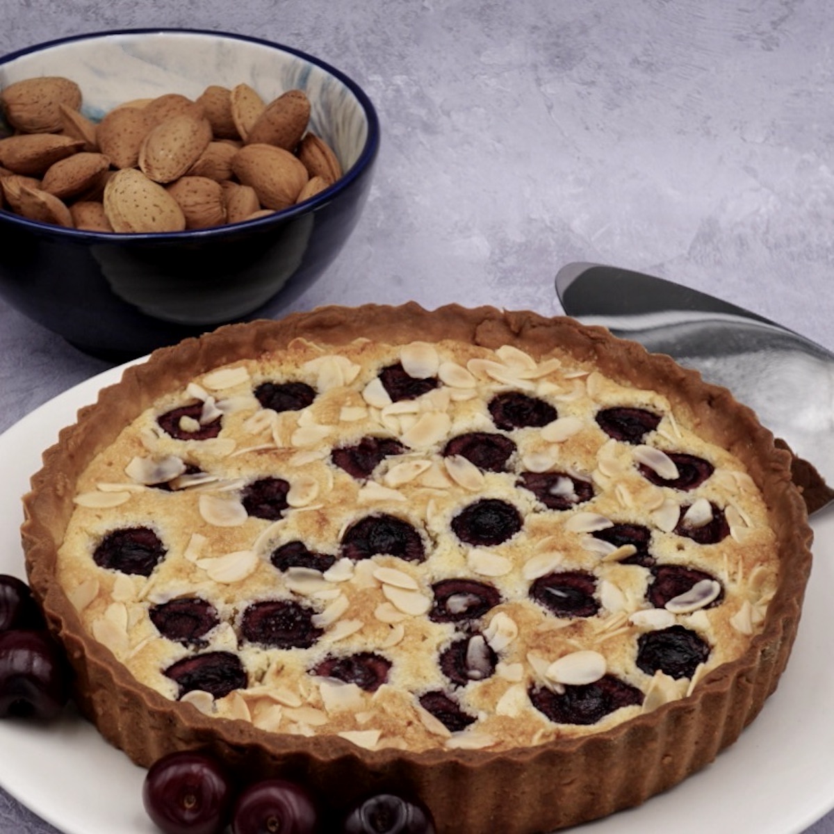 A Bakewell tart on a serving plate next to a bowl of almonds.