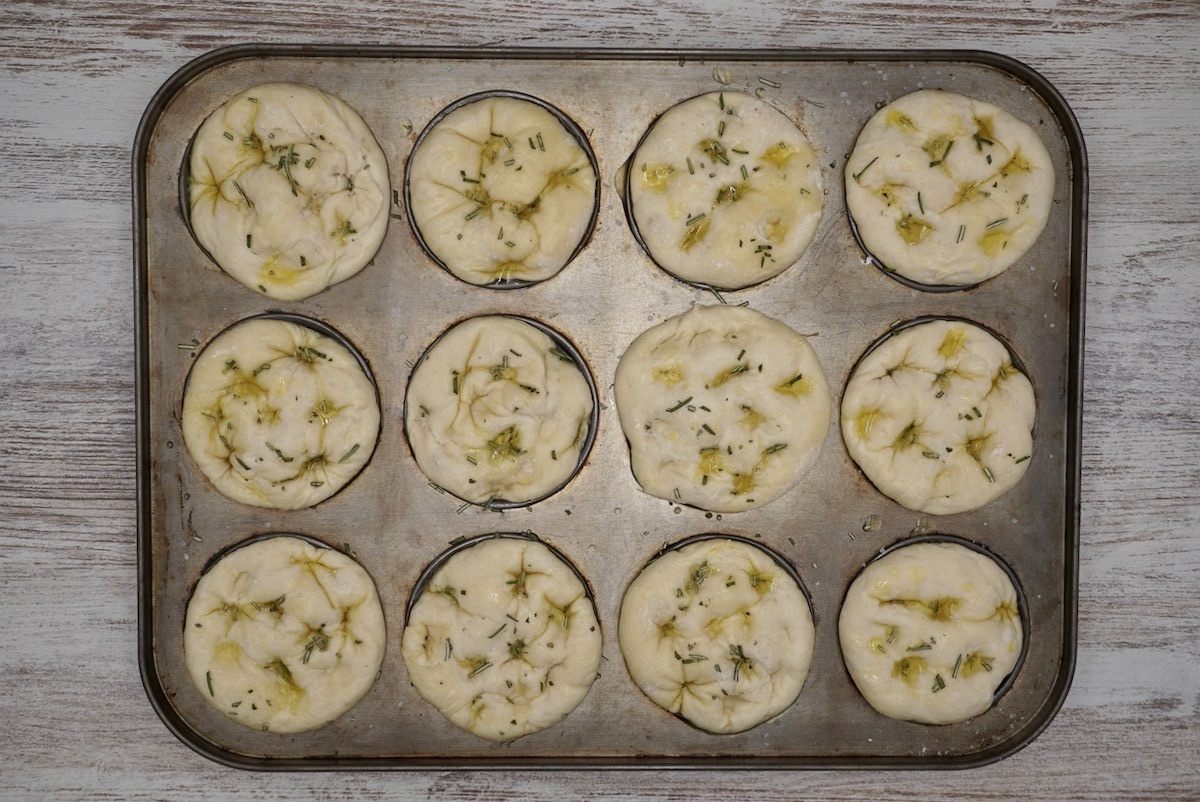 Twlelve unbaked focaccia muffins with olive oil and herbs in a muffin tin.