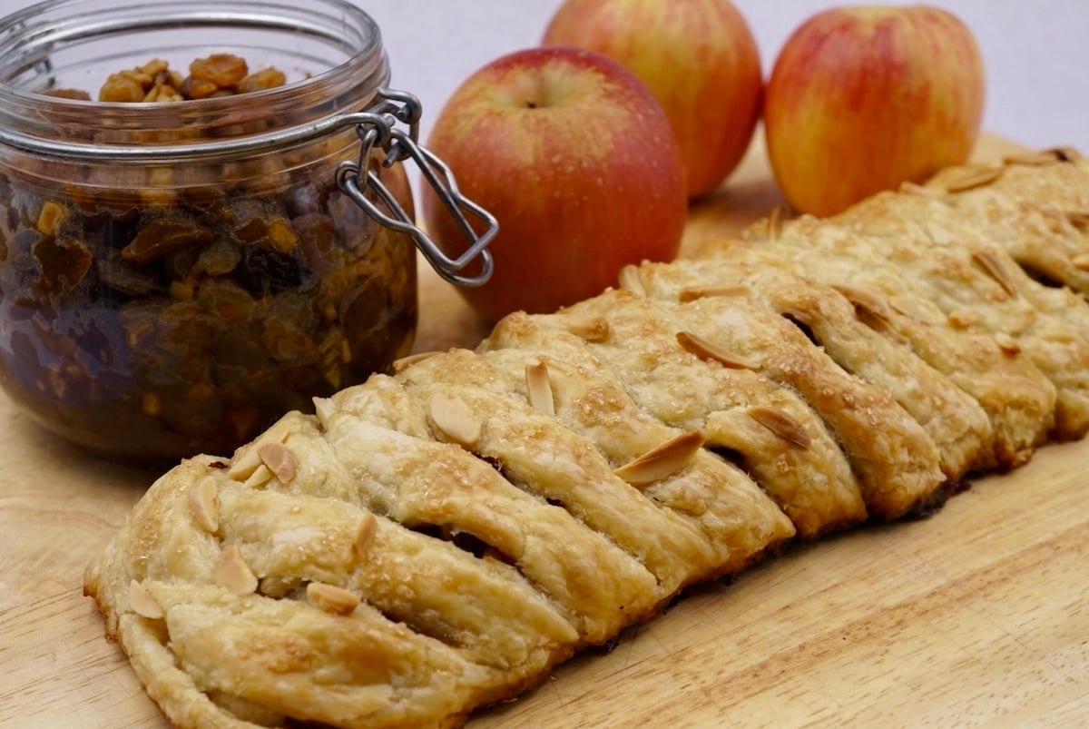 A fruit strudel next to three red apples and a jar of mincemeat.